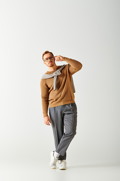 A handsome man in a brown sweater and grey pants striking a stylish pose.