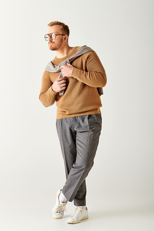 A handsome man strikes a pose in a brown sweater and grey pants.