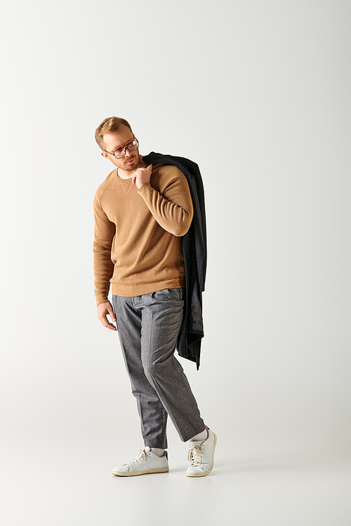 A handsome man poses confidently in a brown sweater and grey pants.