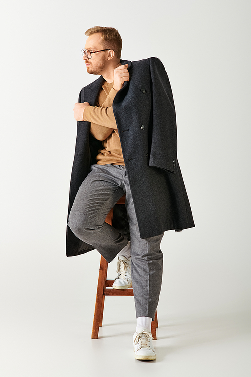 Stylish man seated on stool, exuding charm in chic coat.