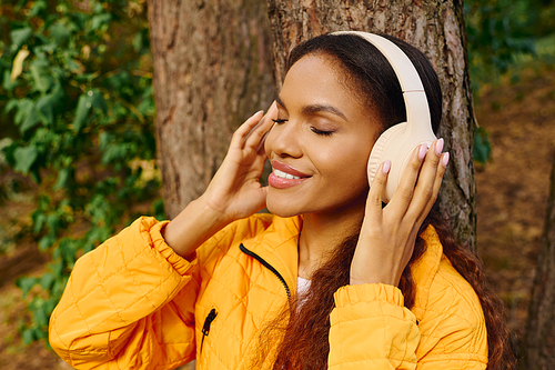 A young African American woman smiles while hiking through an autumn forest, listening to music with joy.