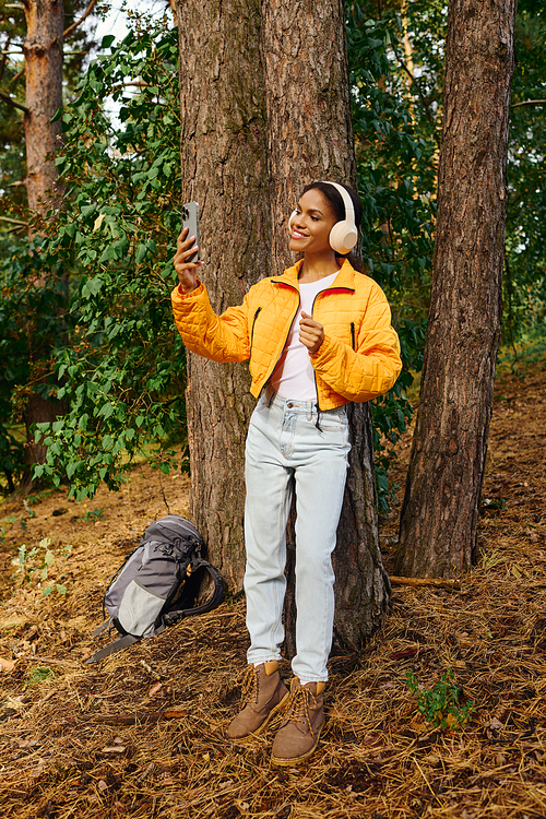 A joyful young woman relaxes against a tree trunk, savoring her hike amidst beautiful autumn foliage.