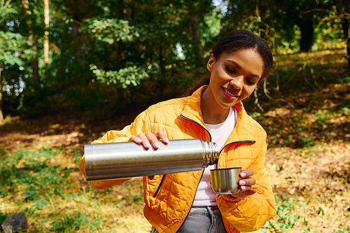 A young woman pours a warm beverage from a thermos, surrounded by colorful autumn foliage while hiking.