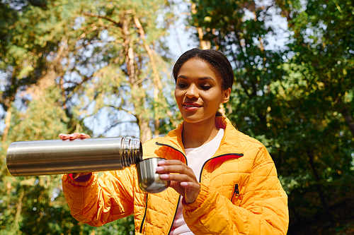 Hiking through a colorful forest, a young woman pours her drink from a thermos, embracing autumn's beauty.