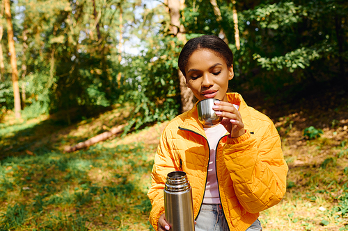 A young woman sips from her cup, soaking in the serene beauty of an autumn forest while hiking.