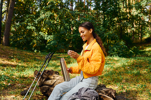 She sits on a fallen log in the forest, savoring a warm beverage surrounded by colorful autumn leaves.