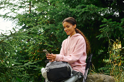 A young African American woman sits on a rock, checking her phone while surrounded by autumn foliage.