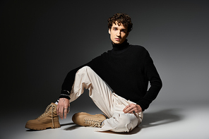 A handsome young man in a black turtleneck elegantly poses against a subtle dark grey backdrop.