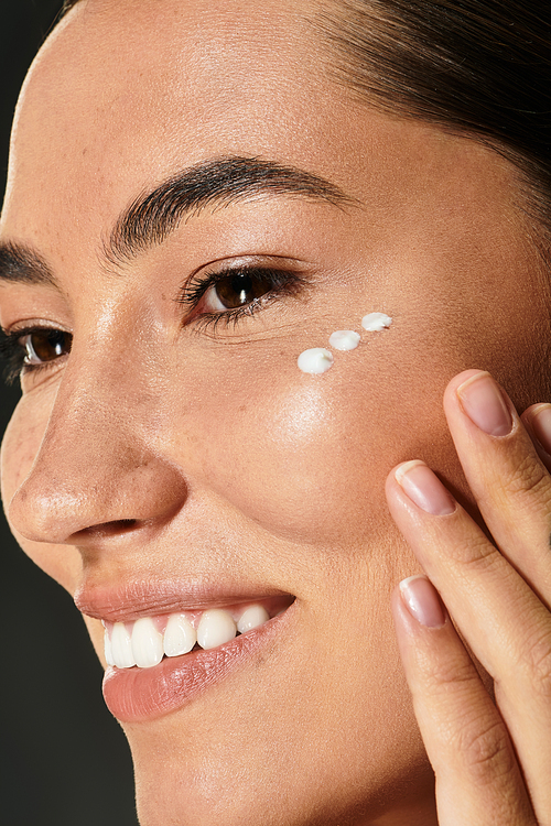 A woman with radiant skin gracefully applies cream to her cheek while smiling.