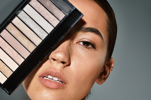 The woman elegantly poses with a makeup palette, highlighting her features in soft light.