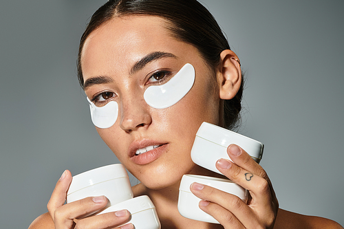 A beautiful woman applies eye masks while holding various skincare products in her hands.