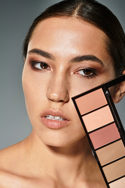 A beautiful woman highlights her features while holding a makeup palette in a stylish environment.