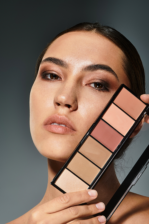 A confident woman elegantly holds a makeup palette, highlighting her flawless skin and artistry.