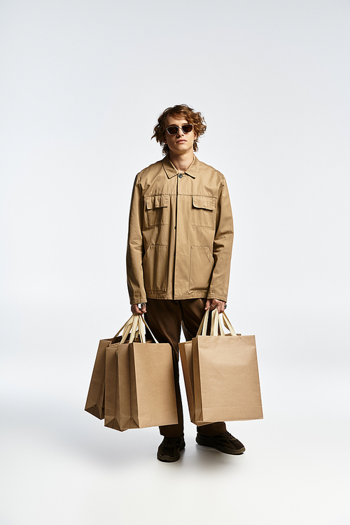 A fashionable young man holds bags while presenting his chic autumn outfit in a studio setting.