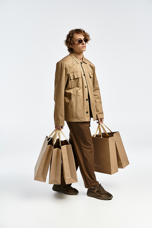 A fashionable young man with wavy hair displays a chic autumn outfit while carrying multiple bags, exuding confidence.