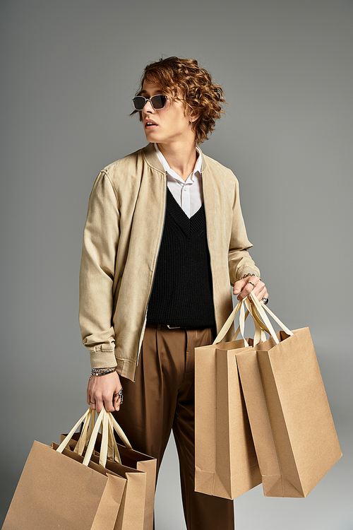 A handsome young man with wavy hair dressed in trendy autumn attire stands confidently in a studio holding bags.