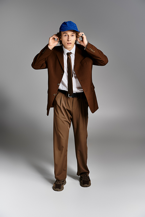 A suave young man stands in a studio, dressed in a brown suit and wearing a blue baseball cap, exuding charm.