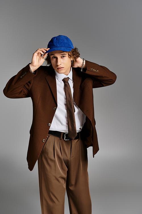 A stylish young man in a brown suit and blue cap adjusts his hat while posing in a contemporary studio.