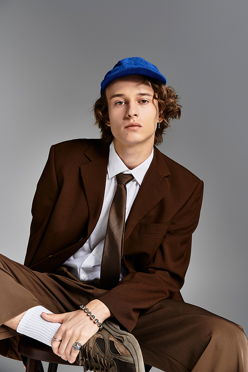 A stylish young man dressed in a brown suit and baseball cap sits confidently in a modern studio.