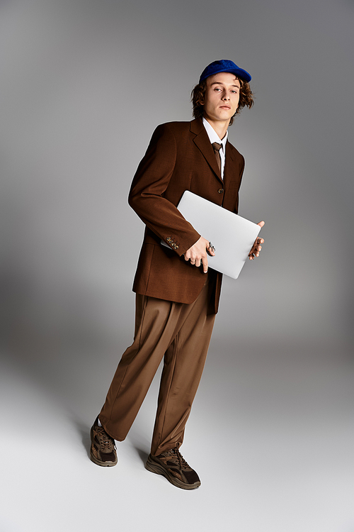 A young man exudes charm in a brown suit and baseball cap while holding a laptop in a chic studio.