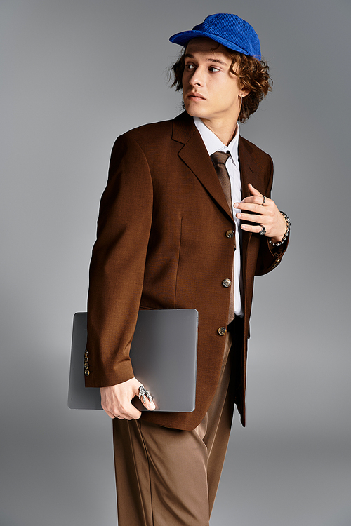 In a modern studio, a handsome young man stands poised, wearing a brown suit and cap, holding a laptop.