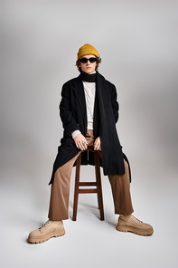 A fashionable young man stands poised in a studio, donning a chic black coat with a playful yellow beanie.