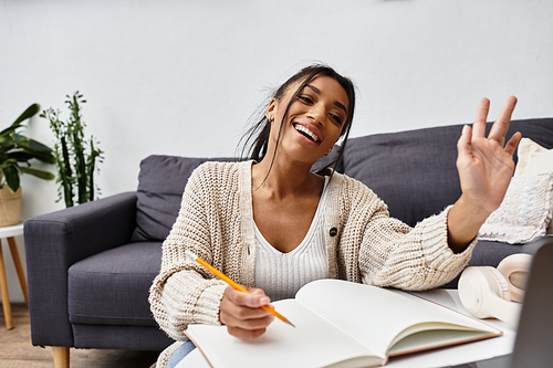 A young woman engages in remote studying at home, smiling and taking notes with enthusiasm.