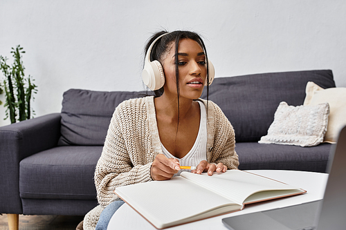 A young woman is focused on her studies at home, taking notes while wearing headphones.