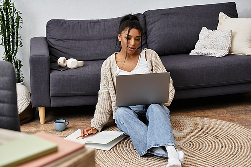 A young woman studies comfortably on the floor, focusing intently on her laptop as she takes notes.