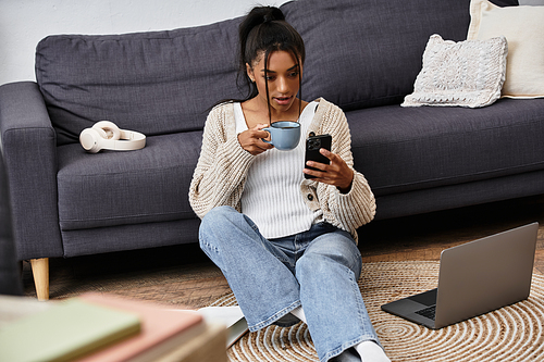 A young woman sits on the floor with a warm drink, focused on her smartphone and studies.