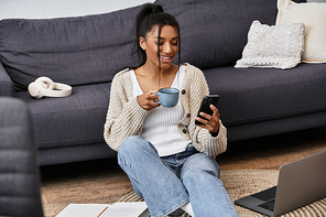 A young woman sits comfortably on the floor, holding a cup of coffee while studying.