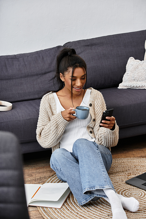 A young woman sits comfortably on the floor, sipping her drink, fully focused on her studies.