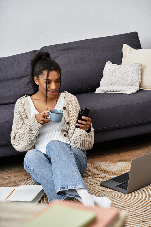 A cheerful woman studies from home, sipping tea and checking her phone while sitting comfortably.