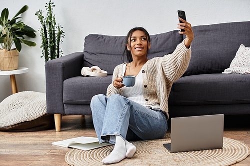 Sitting on the floor, a cheerful woman enjoys coffee and studies while using her laptop.