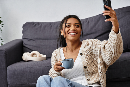 A young woman studies remotely, relaxed on her couch with a cup of coffee and a bright smile.