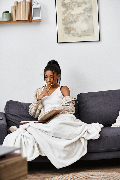 A young woman thoughtfully studies at home, wrapped in a cozy blanket while writing notes.