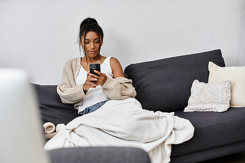 A young woman studies in her cozy living room, focused on her phone and wrapped in a blanket.