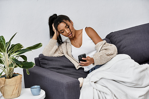 Relaxing on a sofa, a woman engages with her smartphone while studying remotely in comfort.