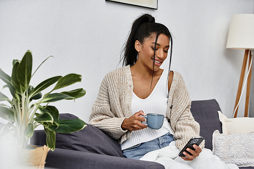 A beautiful woman relaxes at home, studying on her phone while sipping a warm beverage.