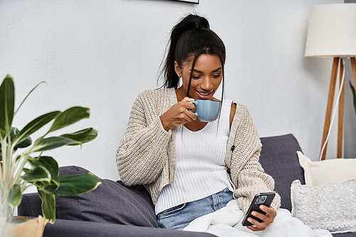 The woman sips her drink while engaged in remote studying on her smartphone at home.