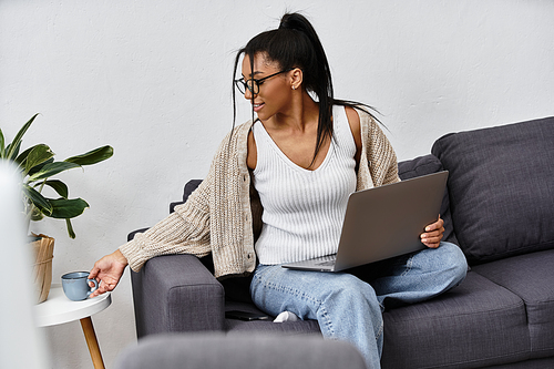 A young woman sitting on a couch, engaged in studying with a laptop and sipping coffee.