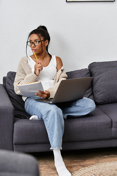 The young woman thoughtfully engages in remote studies, balancing her laptop and notes.