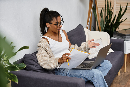 A young woman studies remotely, sitting on a couch surrounded by plants, looking focused.