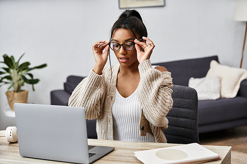 A beautiful woman sits at her desk focused on her laptop, adjusting her glasses.