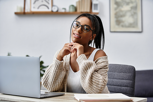 The young woman smiles while studying at home, dressed comfortably and surrounded by books.