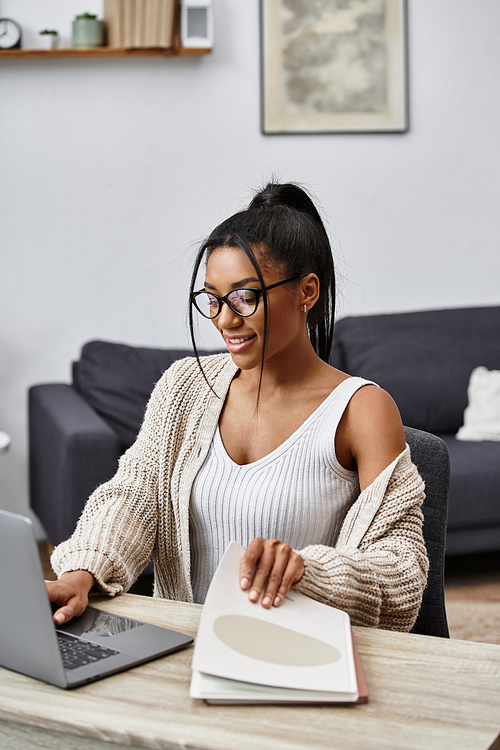 A young woman studies remotely at home, smiling and focused while using her laptop and books.