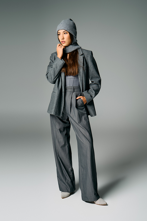 A stylish young woman models her chic gray outfit and cozy knitted hat in a studio.