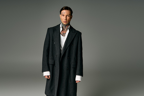 A stylish young man with tattoos poses confidently in a chic coat against a neutral backdrop.