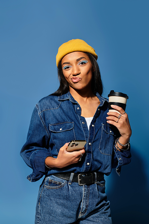 In trendy fall attire, the young woman stylishly poses with coffee against a blue backdrop.