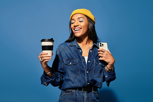 A young woman in warm autumn attire smiles while holding coffee and a smartphone against blue.